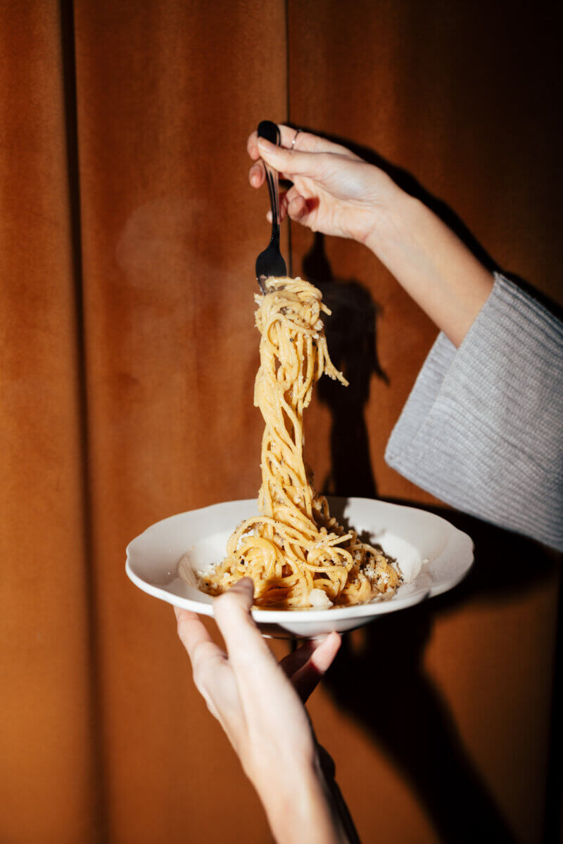 A person twirls spaghetti with a fork over an essential white plate, held in front of a brown curtain background.