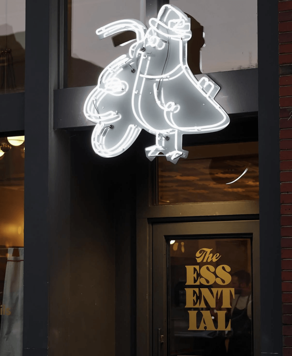 A neon sign of a stylized bird in a hat glows above the door, with “The Essential” written in gold letters—a must-visit spot, especially in the crisp air of fall.