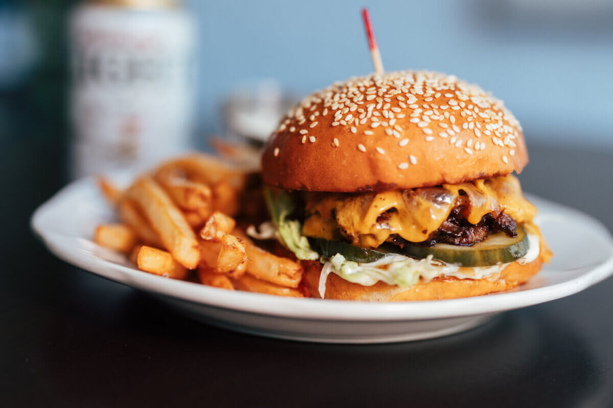 An essential cheeseburger with lettuce, pickles, and sauce on a sesame bun, served with a side of French fries on a white plate—perfect for your next visit this fall.