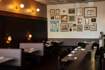 Restaurant interior with empty booths and tables, wall-mounted lamps, and a gallery wall of framed artwork and photographs set a warm, inviting scene—perfect for a cozy fall visit to The Essential. White napkins and dishes are arranged neatly on the tables.