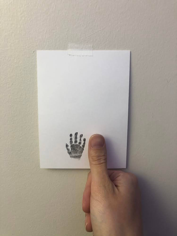 A hand holds a white sheet of paper against a wall; the paper, featuring a black handprint and taped at the top.