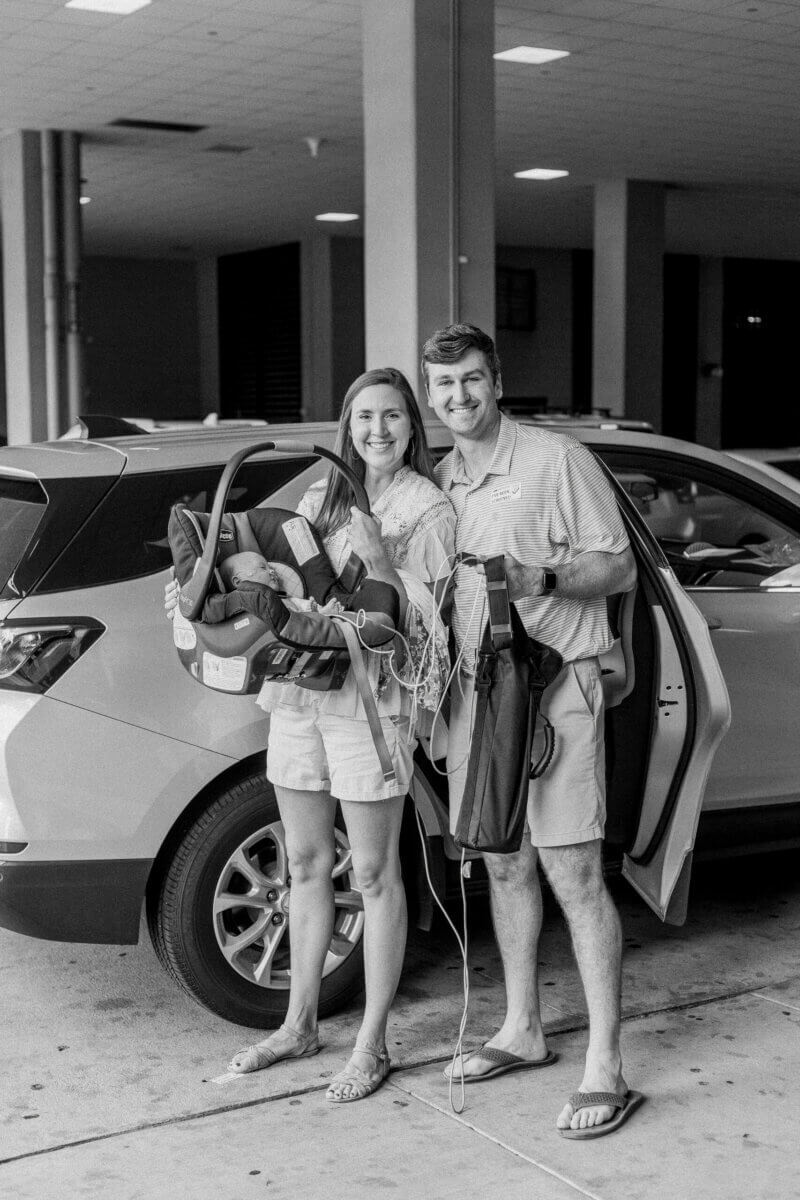 A smiling man and Birmingham mom stand by an open car door holding baby gear and bags, posing for a photo in a covered parking area, capturing a moment of family support after their NICU journey.