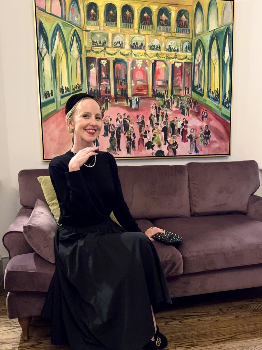 Step into the World of BHAM Artist Morgan Jones Johnston - 2 Morgan Johnston in a black dress sits on a purple sofa, smiling and holding a beaded clutch. A colorful painting of a ballroom scene hangs on the wall behind her.