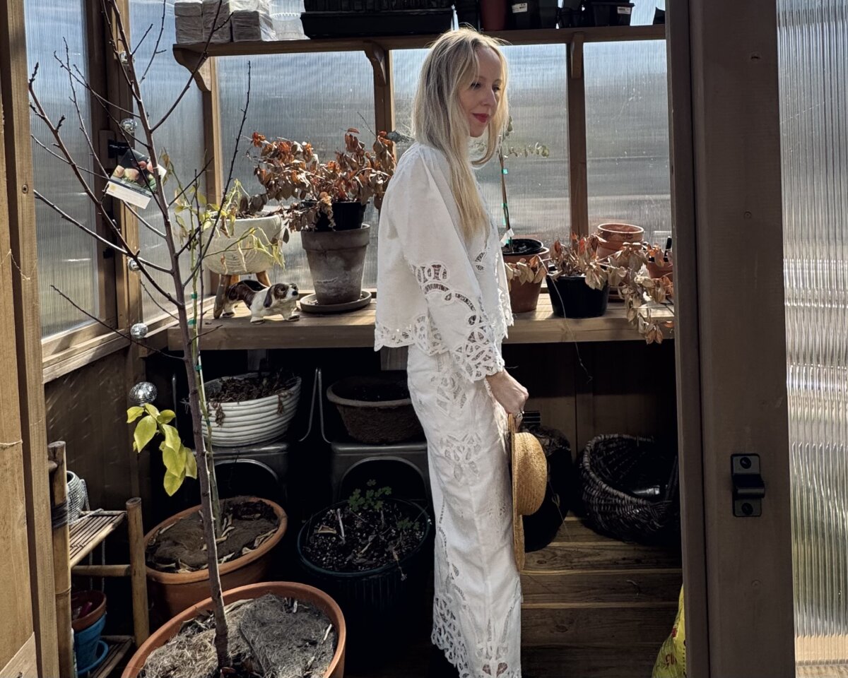 Step into the World of BHAM Artist Morgan Jones Johnston - 7 Morgan Johnston in a white lace dress stands in a sunlit greenhouse, surrounded by potted plants, pots, and gardening tools on wooden shelves.