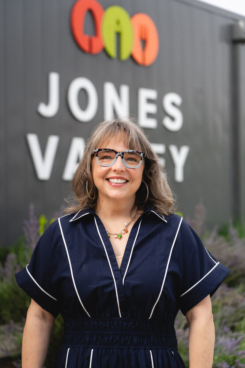 A woman with gray hair and glasses smiles in front of a building with a “JONES VALLEY” sign and a colorful logo above it, highlighting the vibrant spirit of this teaching farm in Birmingham.