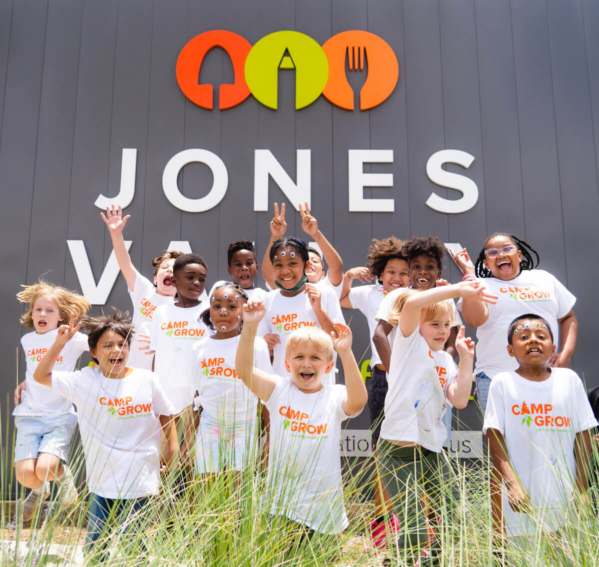 A group of children in “Camp Grow” shirts pose and smile in front of a Jones Valley sign with garden-themed icons above it, celebrating their day at the Birmingham teaching farm.
