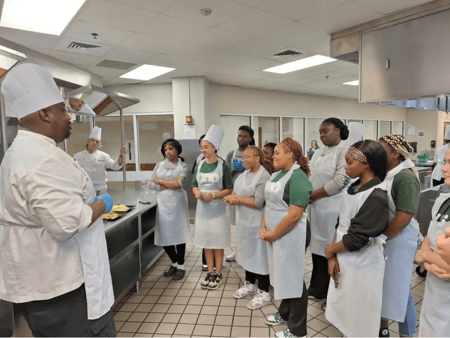 A chef in a white hat instructs a group of students wearing aprons and chef hats in a commercial kitchen.