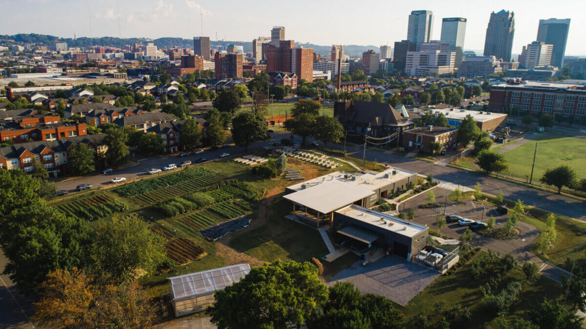 Aerial view of the Birmingham teaching farm with greenhouses and fields, surrounded by buildings and city skyline in the background under a clear sky.