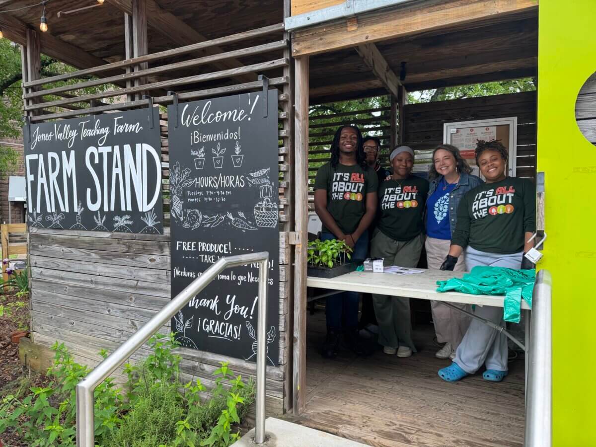 Four people stand behind a wooden farm stand with a chalkboard sign that reads “Welcome! Farm Stand,” surrounded by plants and produce, at an outdoor teaching farm.