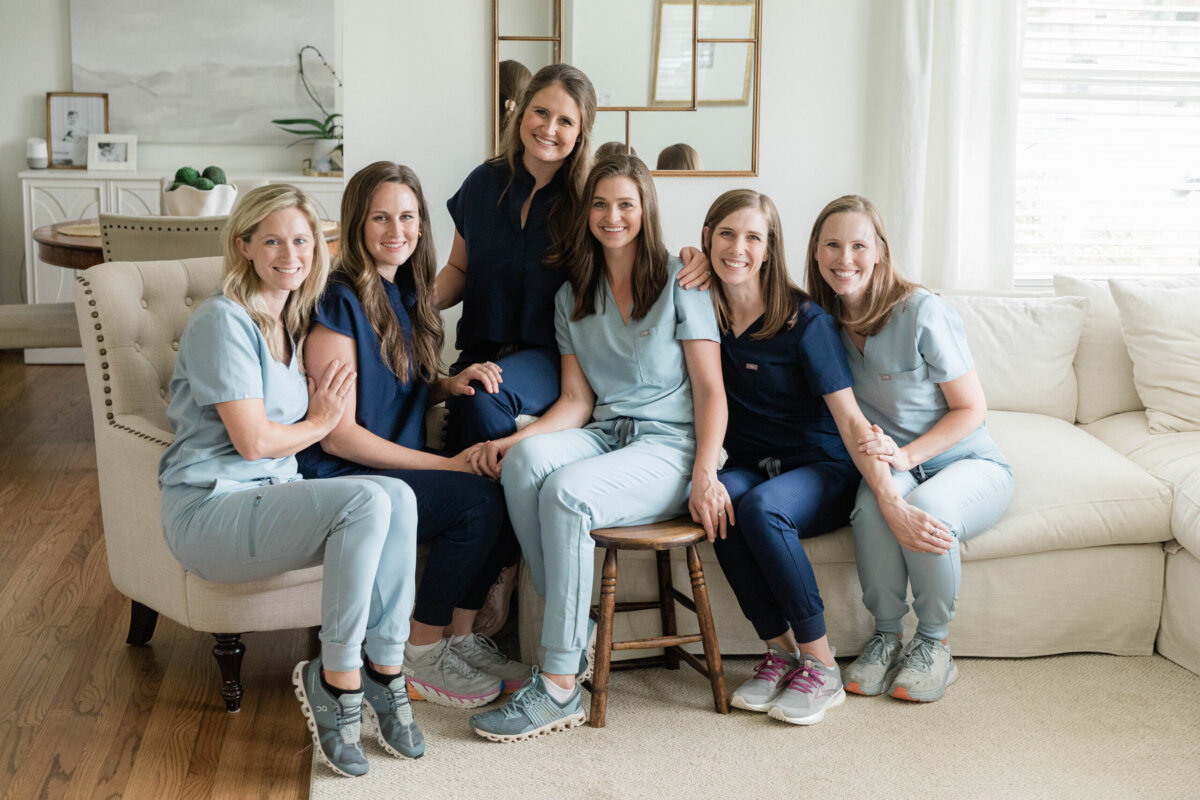 She’s Working to Make Breastfeeding Easier for BHAM Moms - 4 Six women in medical scrubs, all on staff at Birmingham Breastfeeding, sit together on a sofa and a stool in a bright, modern living room in Birmingham, smiling at the camera.