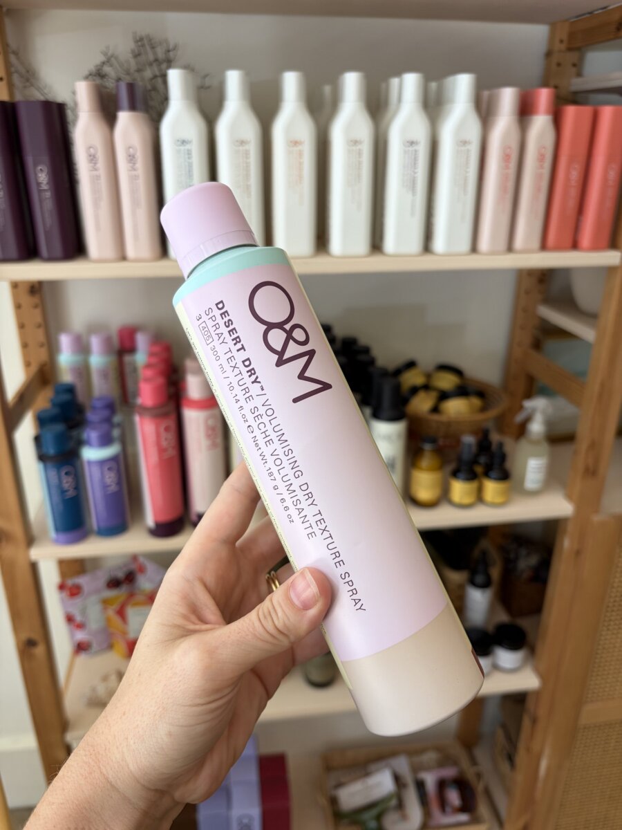 A hand holds a bottle of O&M Desert Dry Volumising Texture Spray in front of shelves stocked with various hair and beauty products, offering September Inspiration for those perfect Fall Football Weekends.