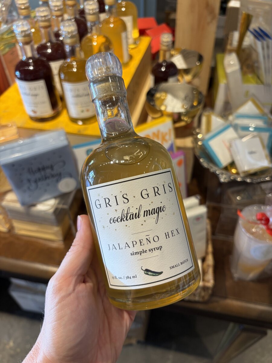 A hand holds a bottle labeled “Gris Gris Cocktail Magic Jalapeño Hex Simple Syrup” in a store with various bottles and products on display in the background, offering perfect September inspiration for crafting unique drinks during fall football weekends.