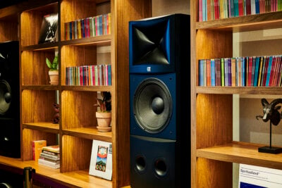 Wooden shelves filled with colorful CD cases, books, and potted plants surround a large black speaker, evoking the cozy atmosphere of Birmingham’s Listening Lounges. A framed poster and decorative items sit on the lower shelf.