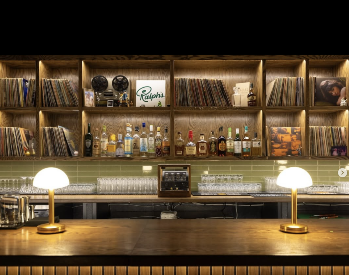 Vinyl, Cocktails & Good Vibes: Birmingham’s New Listening Lounges - 7 A bar with bottles of liquor, glassware, and two lit lamps on the counter. Shelves behind display vinyl records, books, and decorative items, creating the inviting atmosphere of Birmingham’s renowned listening lounges.