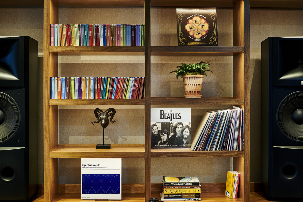 Vinyl, Cocktails & Good Vibes: Birmingham’s New Listening Lounges - 4 Wooden shelves with colorful CDs, vinyl records, a Beatles album, a potted plant, a decorative sculpture, and large black speakers on either side evoke the cozy charm of Birmingham listening lounges.