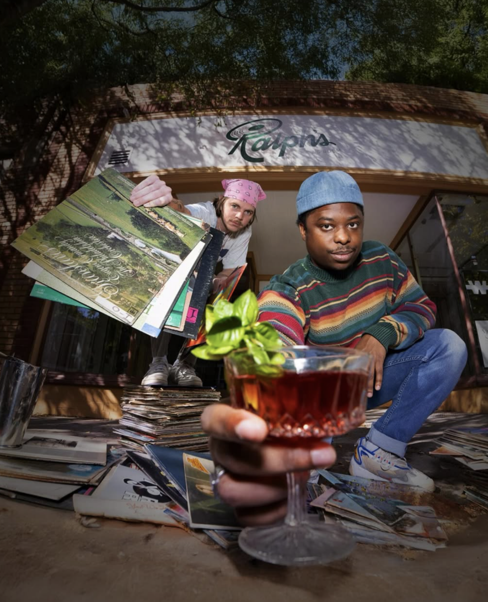 Vinyl, Cocktails & Good Vibes: Birmingham’s New Listening Lounges - 6 Two people pose outside a shop in Birmingham holding vinyl records; one hands a drink with garnish toward the camera. Records are scattered on the ground.