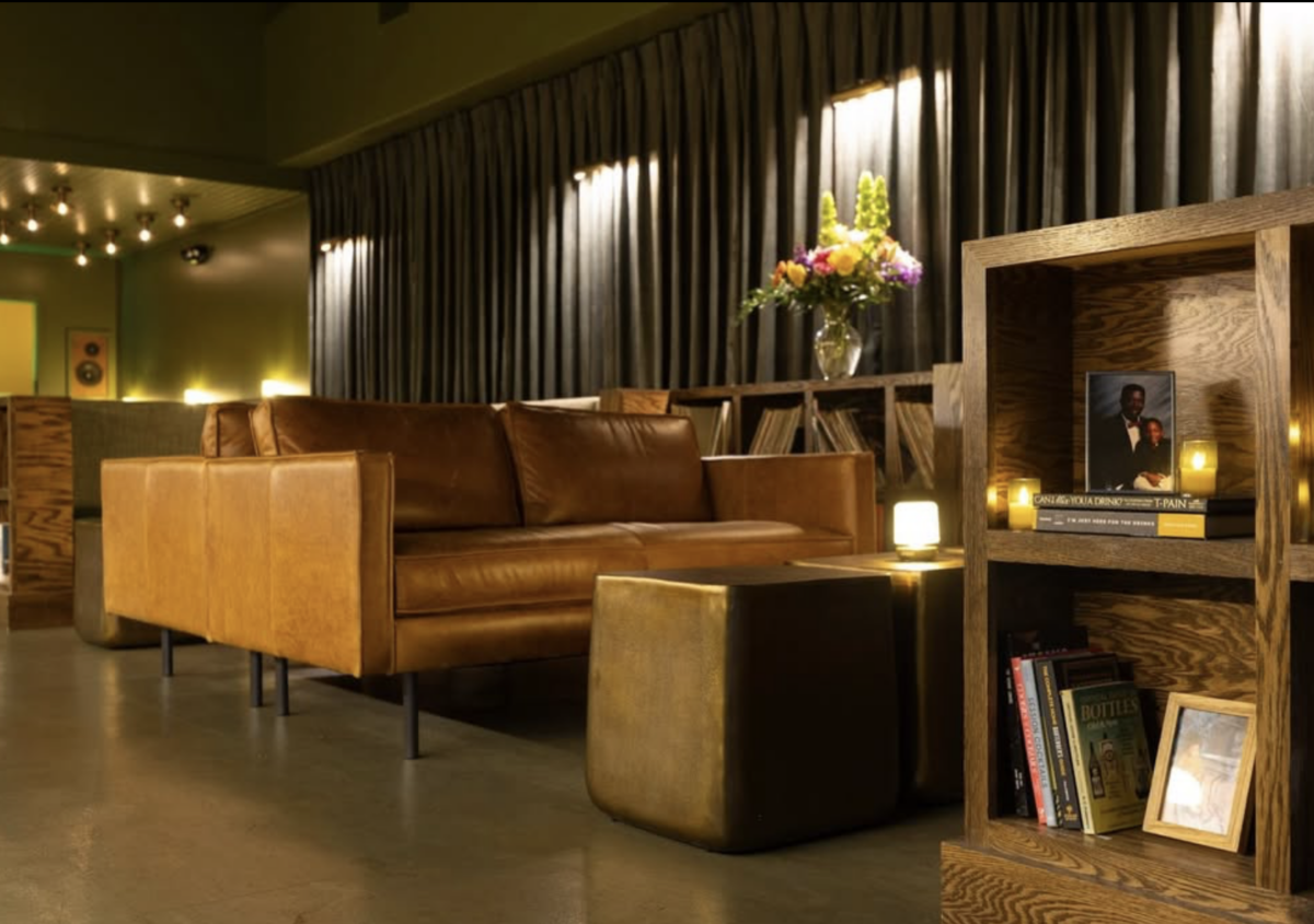 Vinyl, Cocktails & Good Vibes: Birmingham’s New Listening Lounges - 5 A modern lounge in Birmingham with a brown leather sofa, wooden shelves lined with books, small tables, dim lighting, and a vase of flowers on a side table—perfect for those seeking the relaxed ambiance that Listening Lounges are known for.