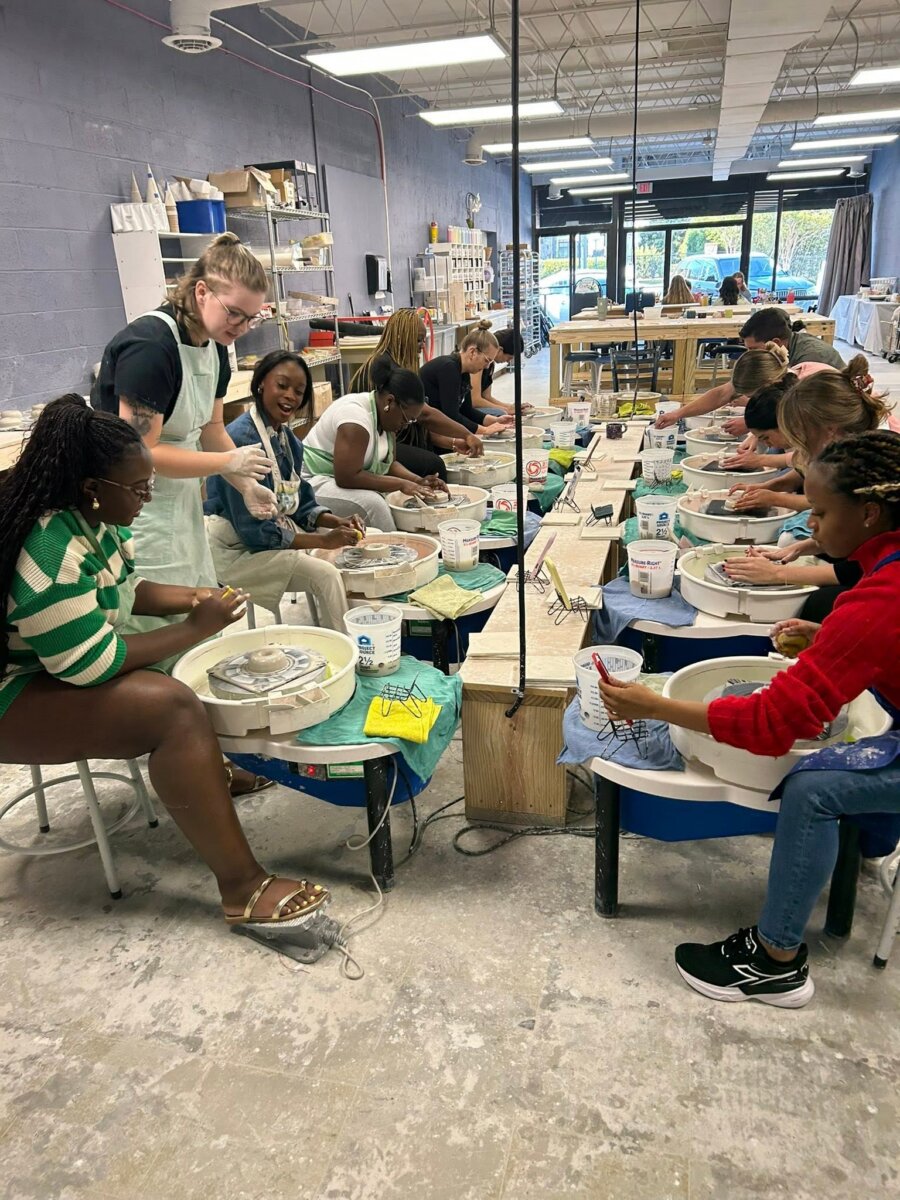 8 Ways to Meet New People in BHAM - 4 A group of people sit at pottery wheels in a ceramics studio at Do It Yourself Crafts. An instructor stands assisting a participant. The room has shelves with supplies and large windows.