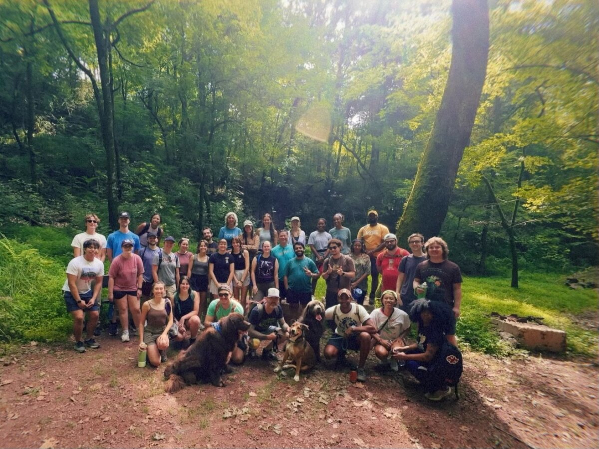 8 Ways to Meet New People in BHAM - 2 A group with Hike Birmingham, along with several dogs, pose for a photo on a dirt path in a wooded area with green trees and sunlight filtering through.