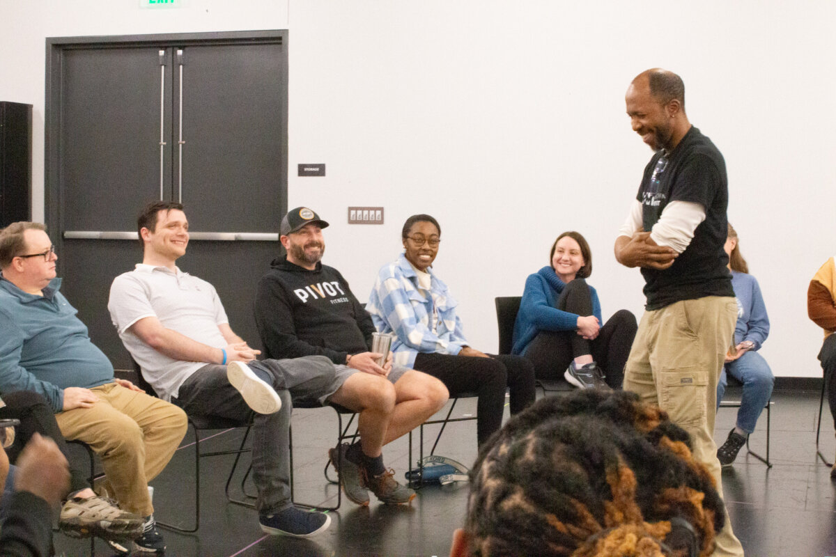 8 Ways to Meet New People in BHAM - 9 A group of people sit in a circle in a bright room in Birmingham, smiling and watching a man standing and speaking at the front, as they enjoy one of many social activities designed to help everyone meet new people.