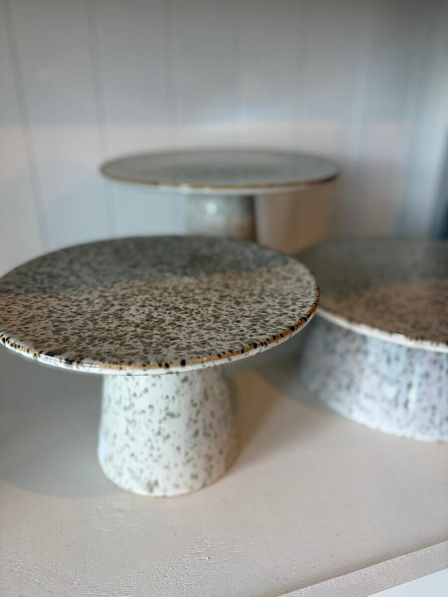 Three speckled ceramic cake stands with wide, round tops and cylindrical bases are displayed on a white shelf against a paneled wall—perfect for showcasing your favorite treats during Fall Football Weekends or as part of your September Inspiration table setting.