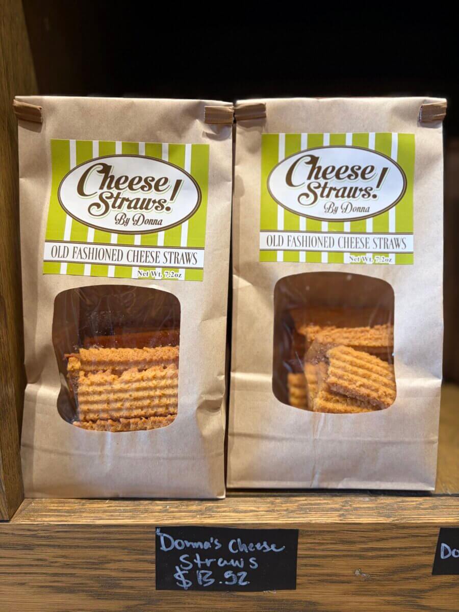 Two brown paper bags labeled “Cheese Straws! By Donna” are displayed on a wooden shelf, each with a clear window showing stacks of cheese straws inside—perfect for Fall Football Weekends. The price tag reads $13.52.