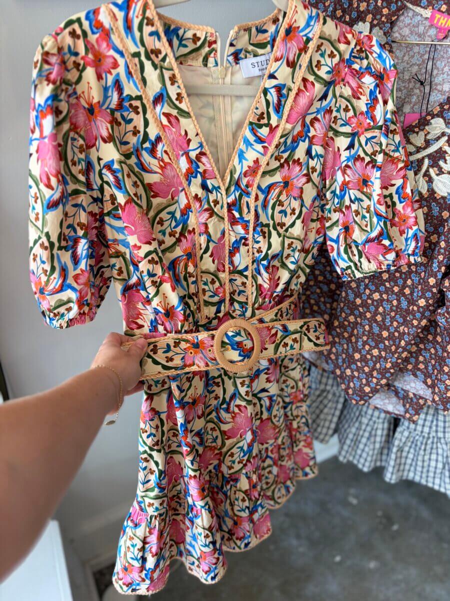A hand holds a short-sleeved floral dress with a matching belt and v-neckline, featuring pink, blue, and green patterns on a cream background—perfect September Inspiration for those crisp Fall Football Weekends. Other dresses hang in the background.