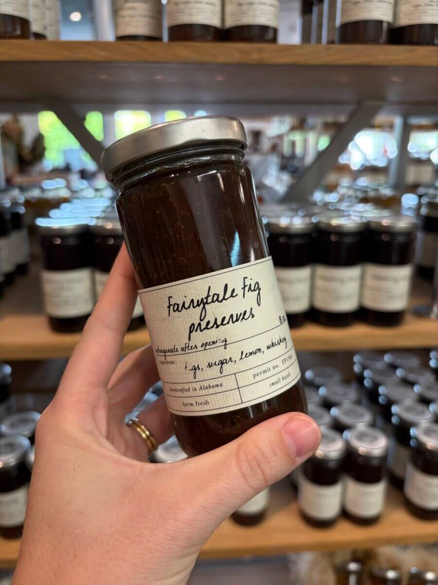 A hand holds a jar labeled “Fairytale Fig Preserves” in front of shelves stocked with similar jars at a store, offering the perfect inspiration for sweet September treats during Fall Football Weekends.