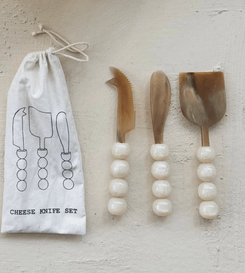 A cheese knife set with three knives featuring pearl-like handles is displayed next to a white drawstring pouch labeled “CHEESE KNIFE SET,” with illustrations of the knives—one of this September’s local must-haves, perfect for fall football weekends and cozy gatherings.