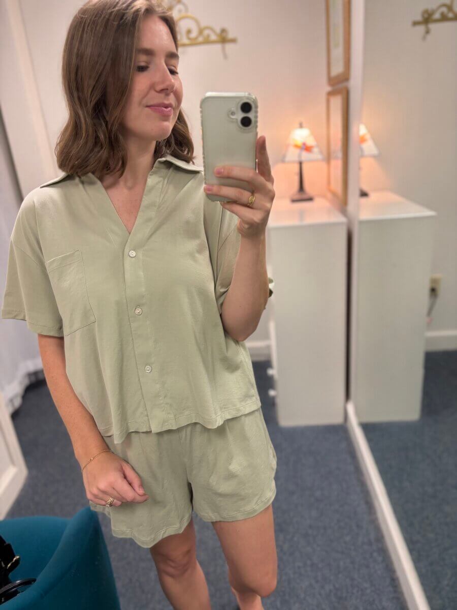 Woman taking a mirror selfie in a light green short-sleeve button-up shirt and matching shorts, serving up September inspiration for cozy Football Weekends, standing in a room with lamps and mirrors.
