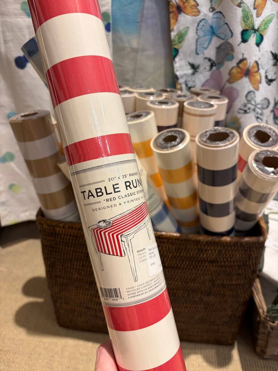 A hand holds a roll of red and white striped table runner in front of a basket filled with various striped table runners in different colors—perfect September inspiration for festive fall football weekends.