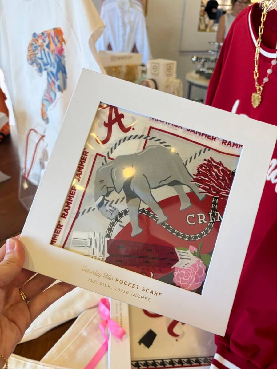 A hand holds a boxed silk pocket scarf featuring an elephant and “Alabama Crimson Tide” themes, displayed in a store with clothing and accessories in the background, offering inspiration for September’s Fall Football Weekends.