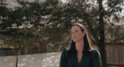 A woman with dark hair wearing a green top stands outdoors, looking to the side, with trees and sunlight in the background, as if thoughtfully reflecting on her fertility journey.