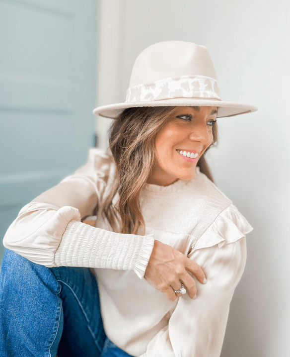This New BHAM Clothing Line Encourages Women to "Wear a Little Joy" - 2 Woman from a Birmingham boutique clothing line wears a beige hat with a patterned band, cream blouse with ruffled shoulder detail, and blue jeans, sitting and smiling by a light wall and blue door.