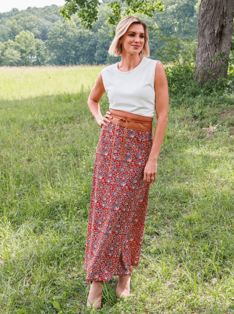 This New BHAM Clothing Line Encourages Women to "Wear a Little Joy" - 3 Woman stands outdoors on grass, wearing a white sleeveless top and a long red patterned skirt with a brown belt—a stylish look from our Birmingham boutique clothing line. Trees and a field are in the background, celebrating women empowerment through fashion.