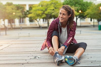Woman sitting outdoors on wooden boards, smiling while tying her running shoe, with earphones and a smartphone beside her—ready to head to the gym, where many women are embracing creatine as part of their fitness routine.