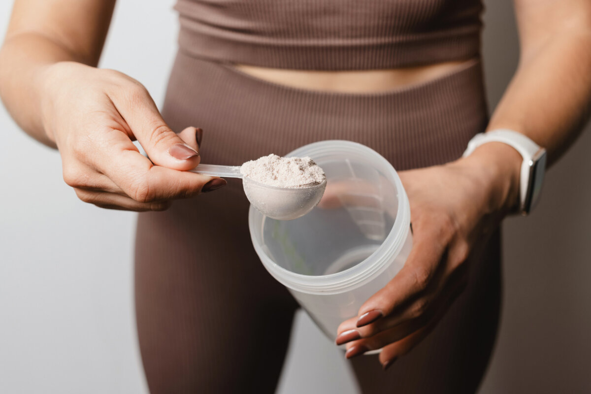 Woman in athletic wear holding a scoop of powder over an open shaker bottle, preparing a drink before heading to the gym.