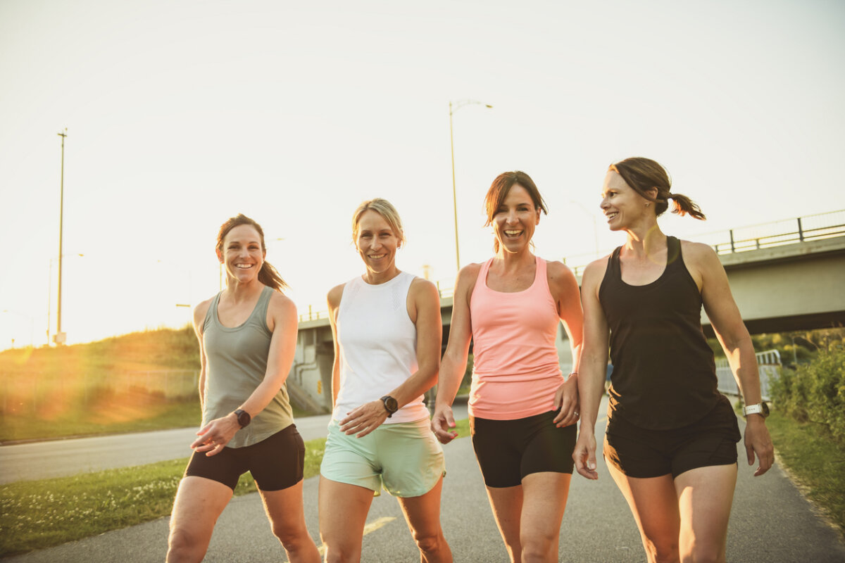 Four women in athletic wear walk together on a paved path outdoors near an overpass, smiling in the sunlight.