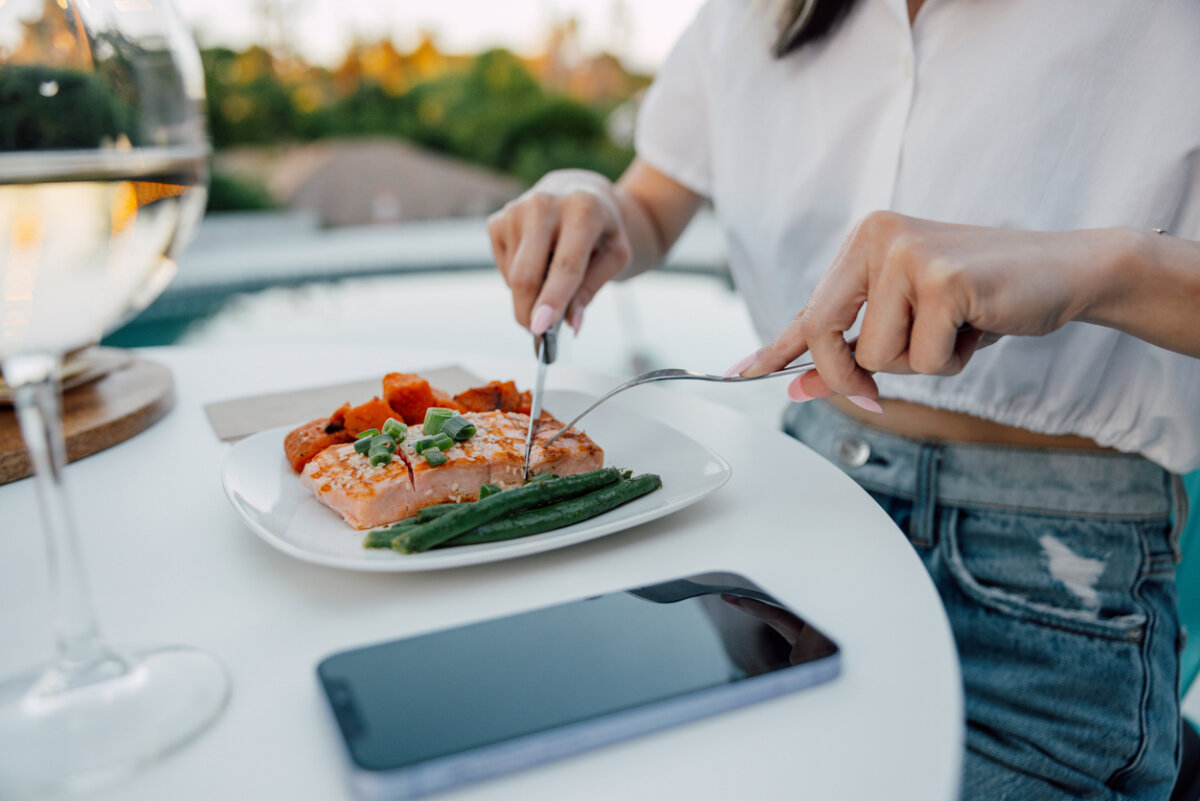 A woman cuts into a salmon fillet with vegetables at an outdoor table.