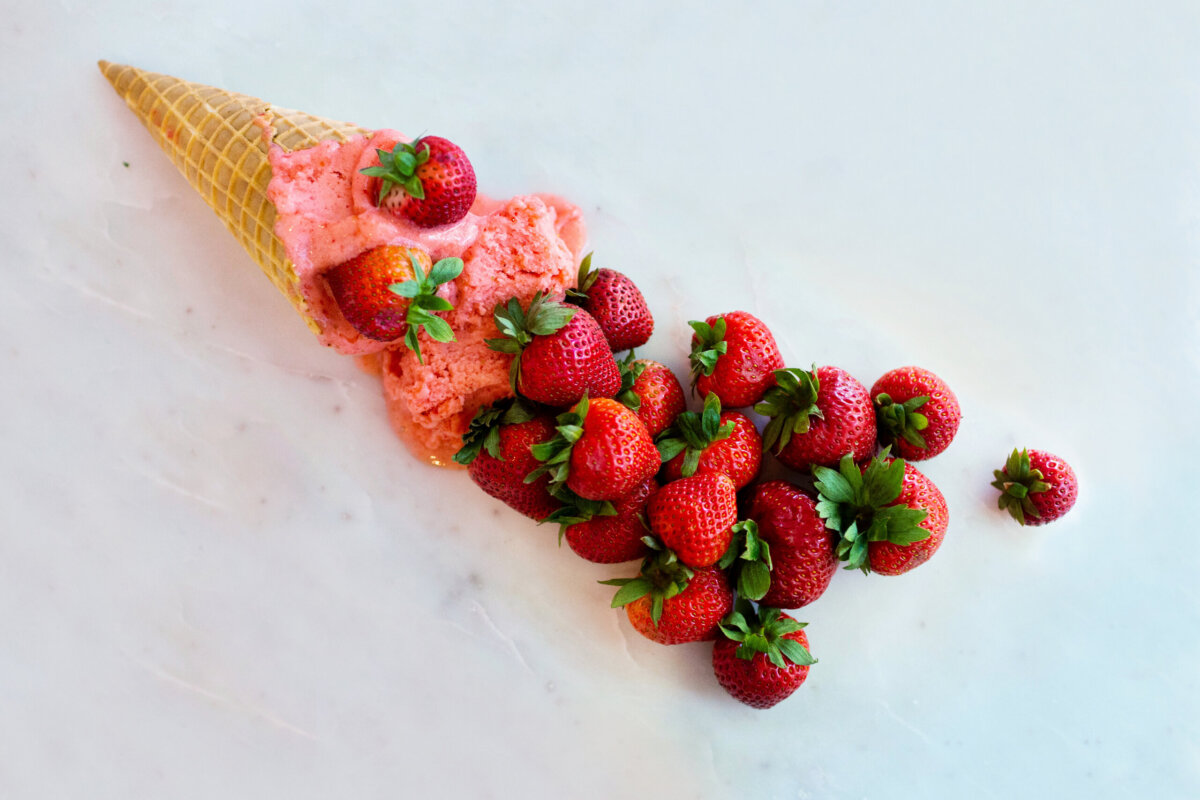 Meet the Art Historian Behind Louisville’s Gelato Gilberto - 4 A waffle cone with strawberry ice cream lies on its side, surrounded by several fresh strawberries scattered next to it on a white surface—a delightful glimpse into the sweet life.
