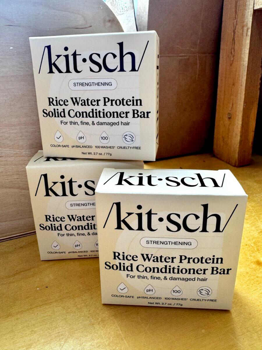 Three boxes of Kitsch Rice Water Protein Solid Conditioner Bar are stacked on a wooden surface, making them true home must haves for anyone focused on hair care. The packaging highlights strengthening benefits for thin, fine, and damaged hair.