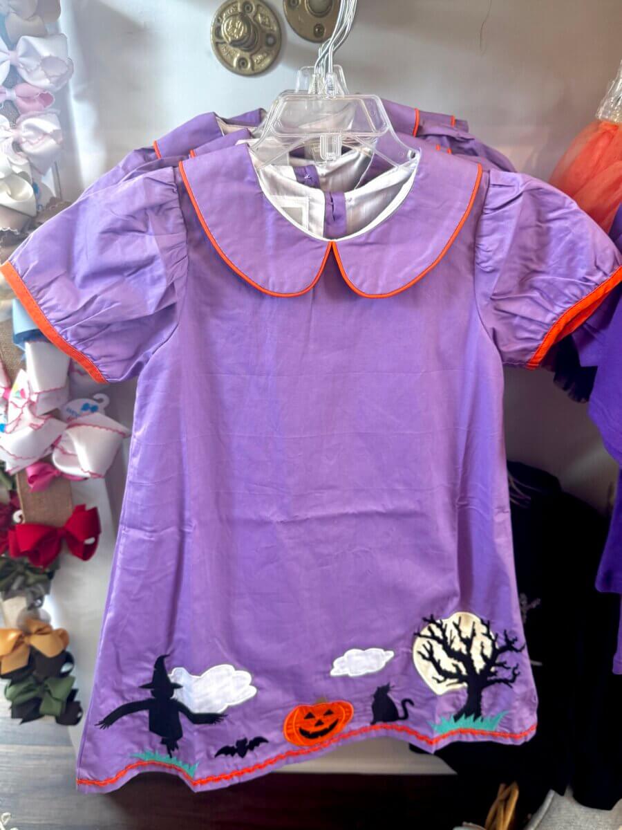 A purple short-sleeve dress with orange trim features Halloween-themed appliqués—including a pumpkin, black cat, tree, and a witch flying on a broom near the hem.