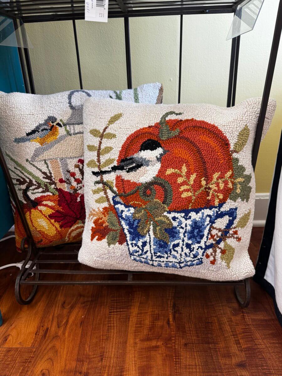 Two decorative throw pillows with bird and fall-themed embroidery rest on a metal rack, one featuring a bird on a pumpkin in a blue teacup, the other with birds among gourds and foliage—a charming Fall Preview of Home Must Haves from Louisville FINDS.
