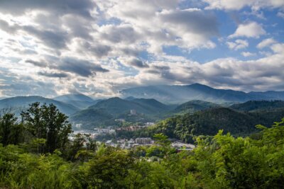 A town sits at the base of forested mountains under a partly cloudy sky, with sunlight streaming through the clouds—inviting visitors to discover hidden gems and charming restaurants nestled throughout Gatlinburg.