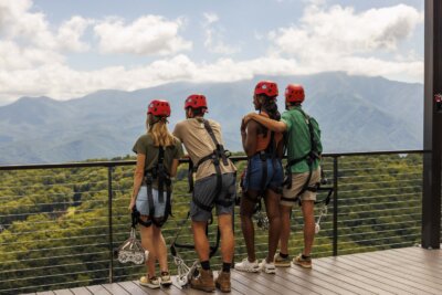 Four people in helmets and harnesses stand on a deck overlooking a forested mountain range under a partly cloudy sky.