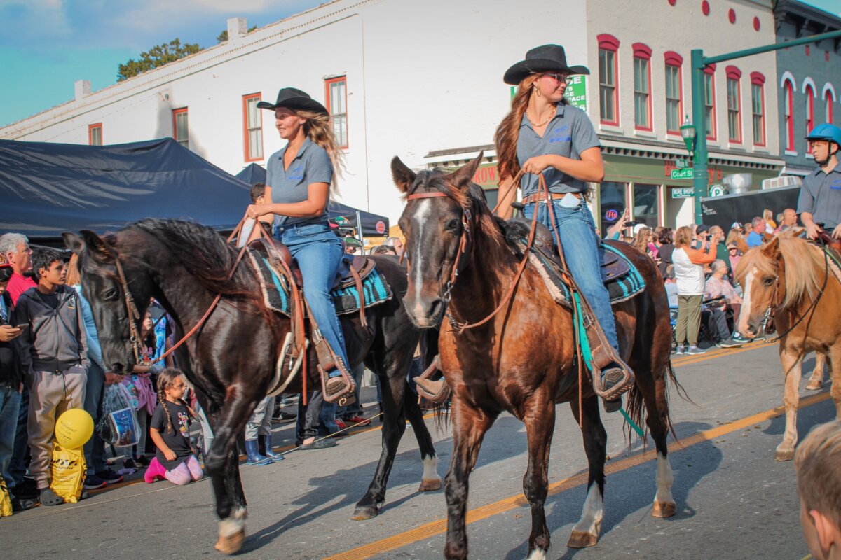 Two women in cowboy hats ride horses in a parade, passing by a crowd on a city street lined with shops—a lively scene reminiscent of beloved Kentucky festivals and small-town celebrations.