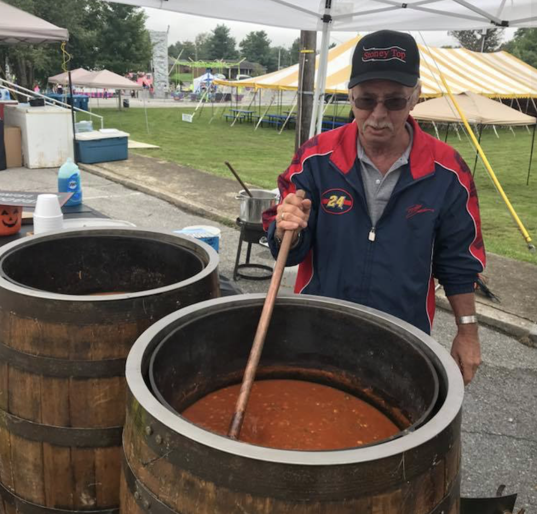 A man in a racing jacket and cap stirs a large wooden barrel filled with stew at an outdoor event under a canopy.