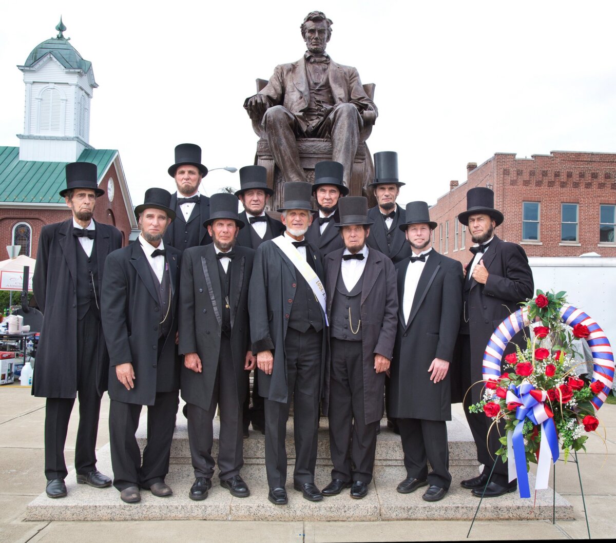 Twelve men in black suits and top hats pose in front of a large Abraham Lincoln statue, with a red, white, and blue wreath on the steps nearby.