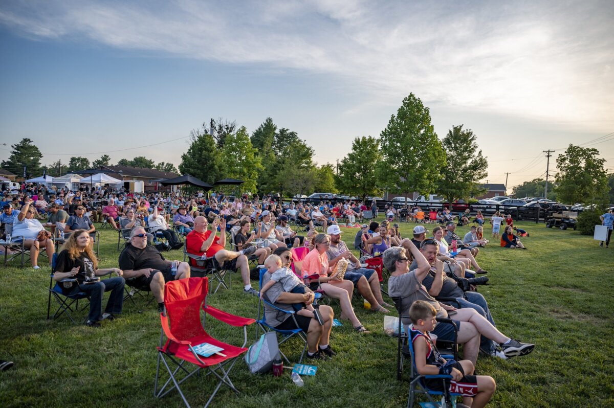 A large group of people sit on lawn chairs and blankets in a grassy park, enjoying one of the many Kentucky festivals as they watch an outdoor event in the early evening.