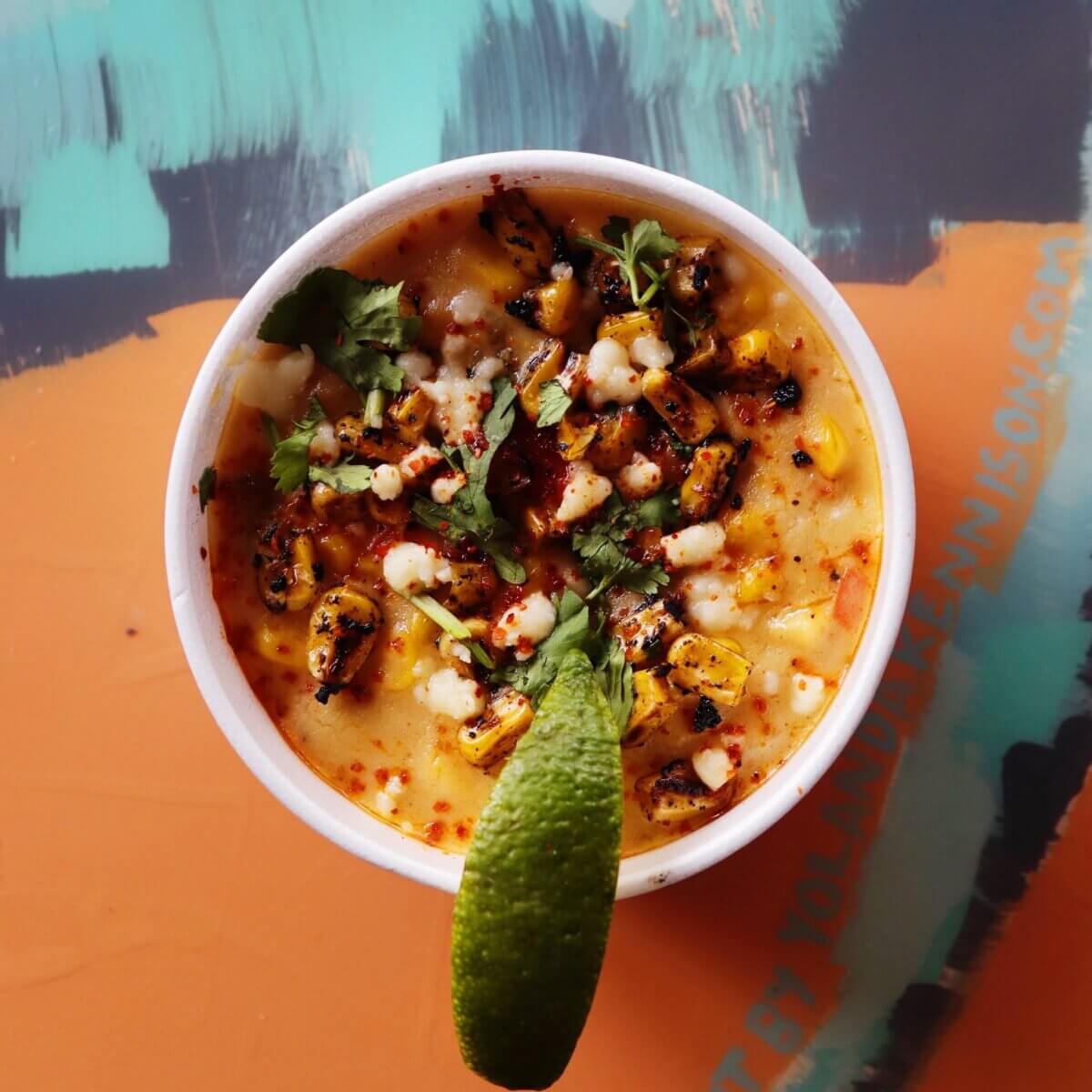 The Best of Lexington, KY: Iconic Stops + Hidden Gems - 6 A white bowl of Mexican street corn topped with cilantro, cheese, chili powder, grilled corn, and a lime wedge, set on a colorful abstract background—perfect for discovering bold flavors in the heart of Lexington, the Horse Capital.
